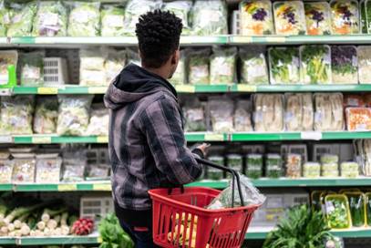 A young man holding a red basket looks at rows of fresh produce displayed on supermarket shelves.